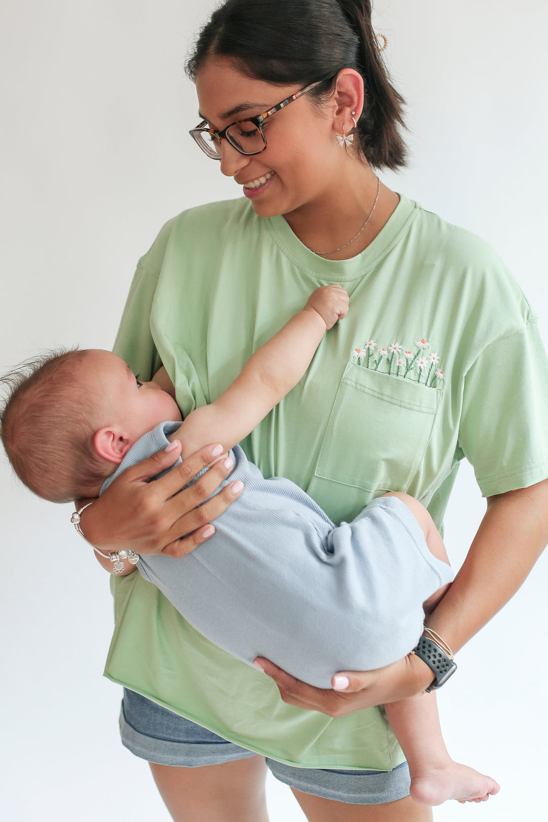 Woman breastfeeding a baby against a plain background. She is wearing an innovative green nursing t-shirt with invisible zipper access to breastfeed.